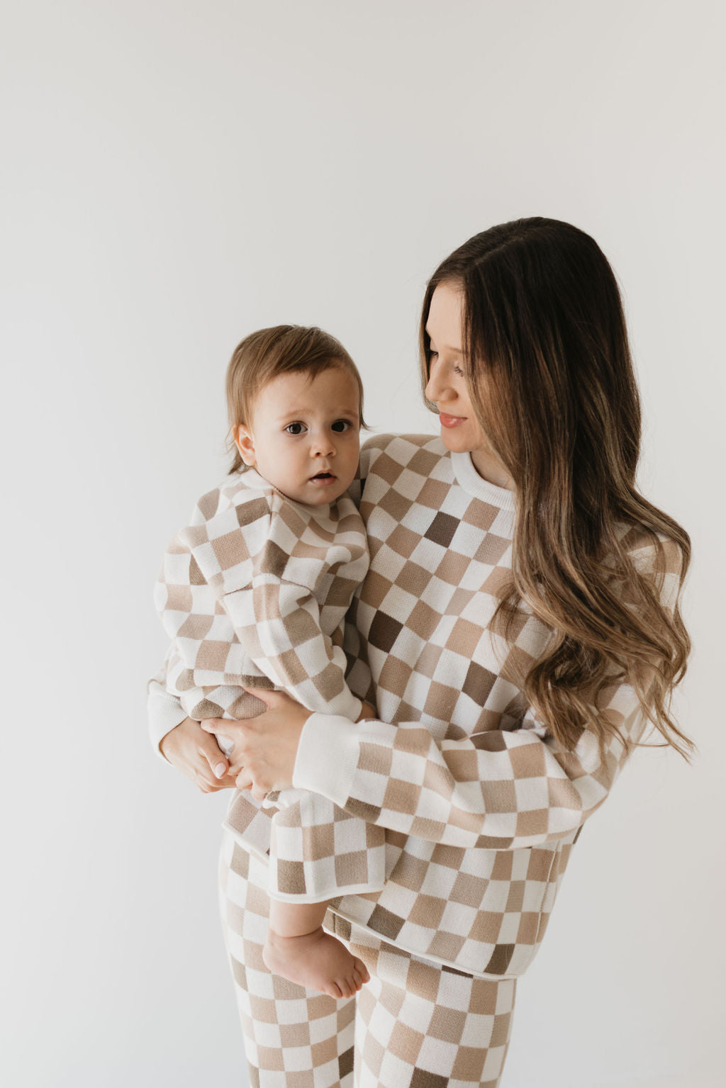 A woman with long hair cradles a baby, both dressed in the "Into the Woods Checkerboard" Knit Pant Set from forever french baby. Their matching outfits are made from 100% cotton and stand out against a plain, light background.