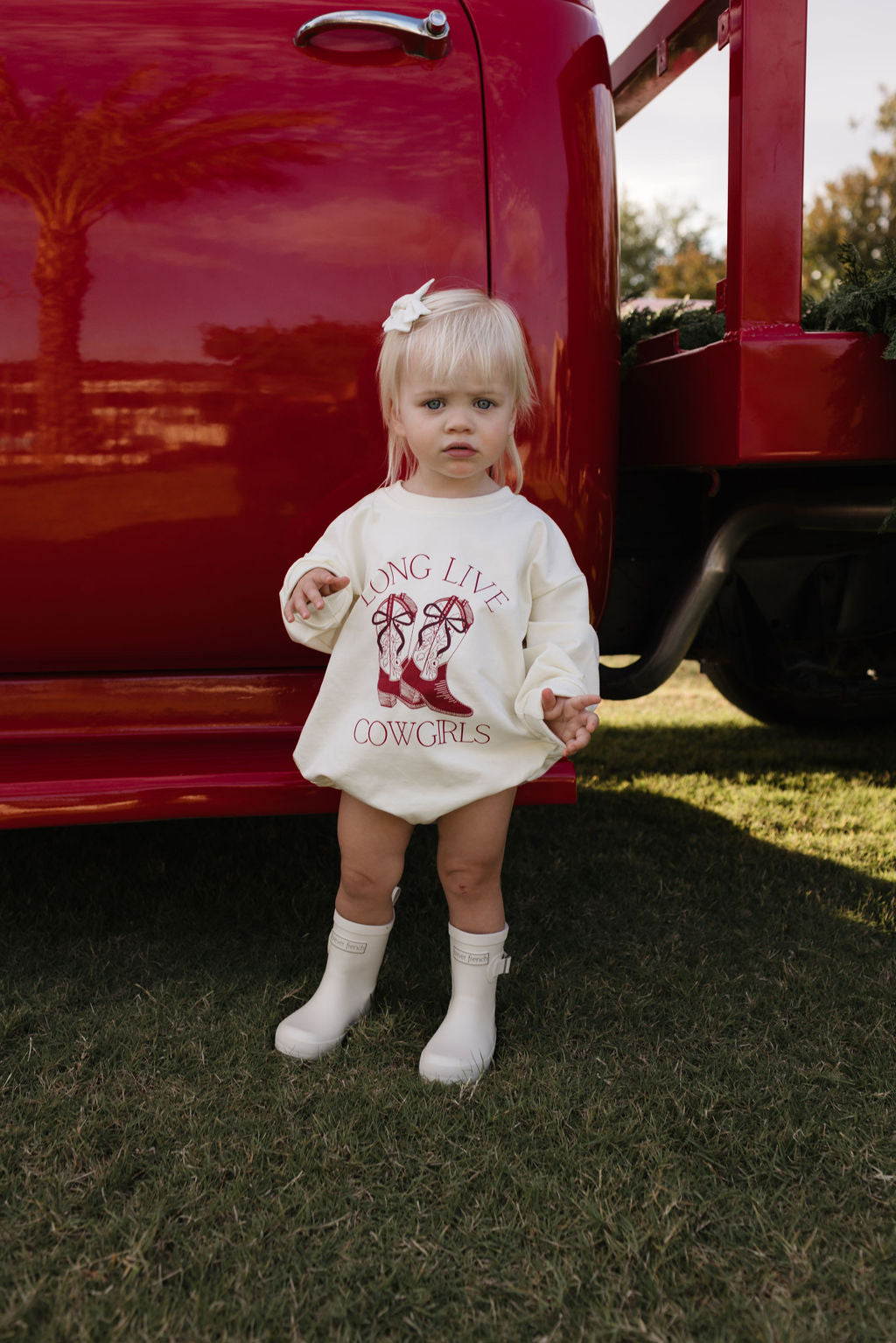 A toddler with blonde hair and a bow stands on grass in front of a red vehicle, dressed in the "Long Sleeve Romper | Long Live Cowgirls" by forever french baby. The cream sweatshirt romper features red text that reads "Long Live Cowgirls" along with images of cowgirl boots. She complements her outfit perfectly for the cooler months with white boots.