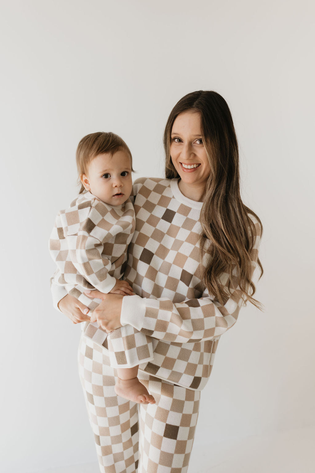 A woman with long hair smiles as she holds a toddler, both adorned in matching "Into the Woods Checkerboard" knit pant sets by forever french baby. These 100% cotton outfits contribute to a cozy ambiance against the simple white background, enhancing the image's clean and minimalistic appearance.