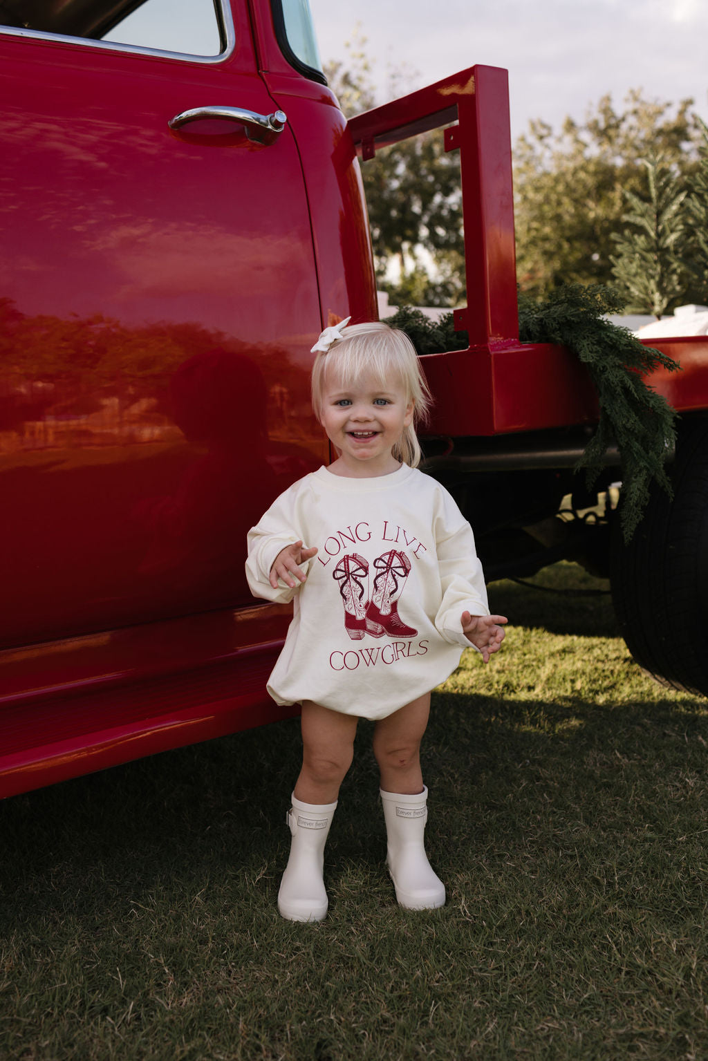 A young child with blonde hair stands smiling in front of a red truck on grass, embracing the cooler months in a cozy Long Sleeve Romper from forever french baby, featuring the print "Long Live Cowgirls." White boots complete the look as greenery adorns the truck and trees sway gently in the background.