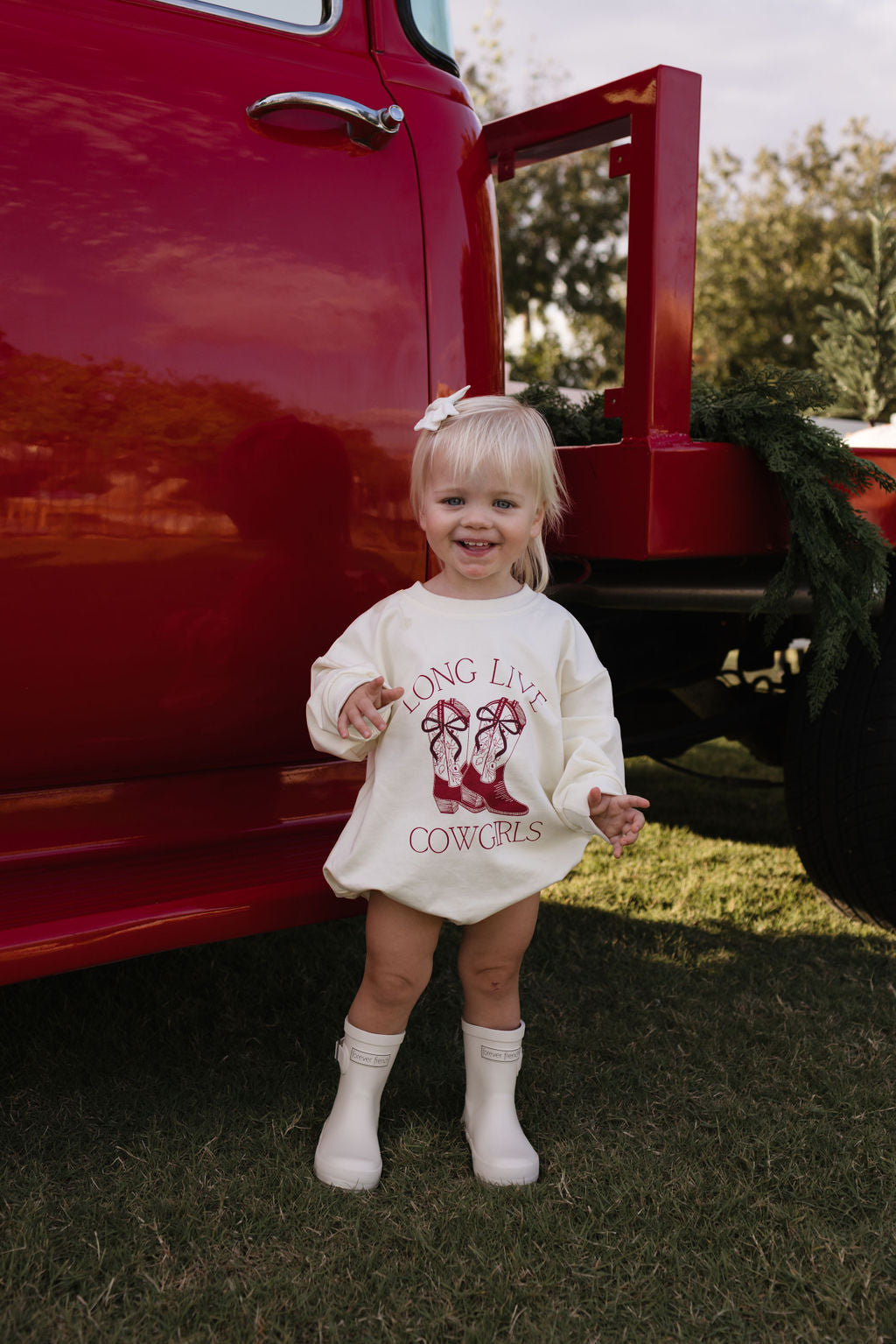 A toddler with light hair stands in front of a red truck, wearing the "Long Sleeve Romper | Long Live Cowgirls" by forever french baby, paired with white boots. Perfect for cooler months, the scene's backdrop features grass and trees under a clear sky.