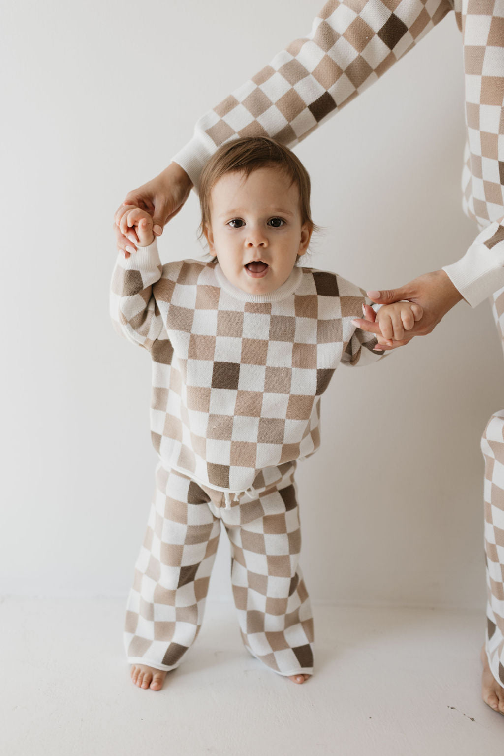 A toddler dressed in the "Into the Woods Checkerboard" knit pant set by forever french baby stands holding hands with an adult. Made from 100% cotton, the beige and white checkered outfit displays a delightful pattern. The toddler looks to be taking a step forward, while the adult is wearing a matching oversized top. The background is plain white.