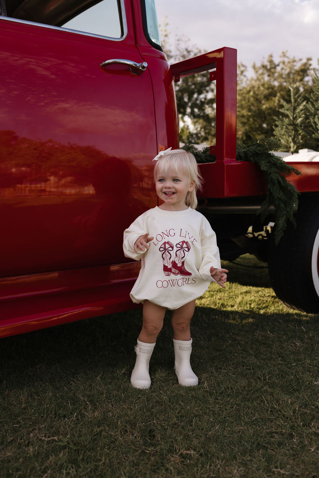 A smiling toddler stands on the grass in front of a red truck, wearing the cream-colored "Long Sleeve Romper | Long Live Cowgirls" by forever french baby. The outfit suits cooler months and is perfectly paired with white boots and a hair bow, completing the western style accentuated by greenery visible on the truck.