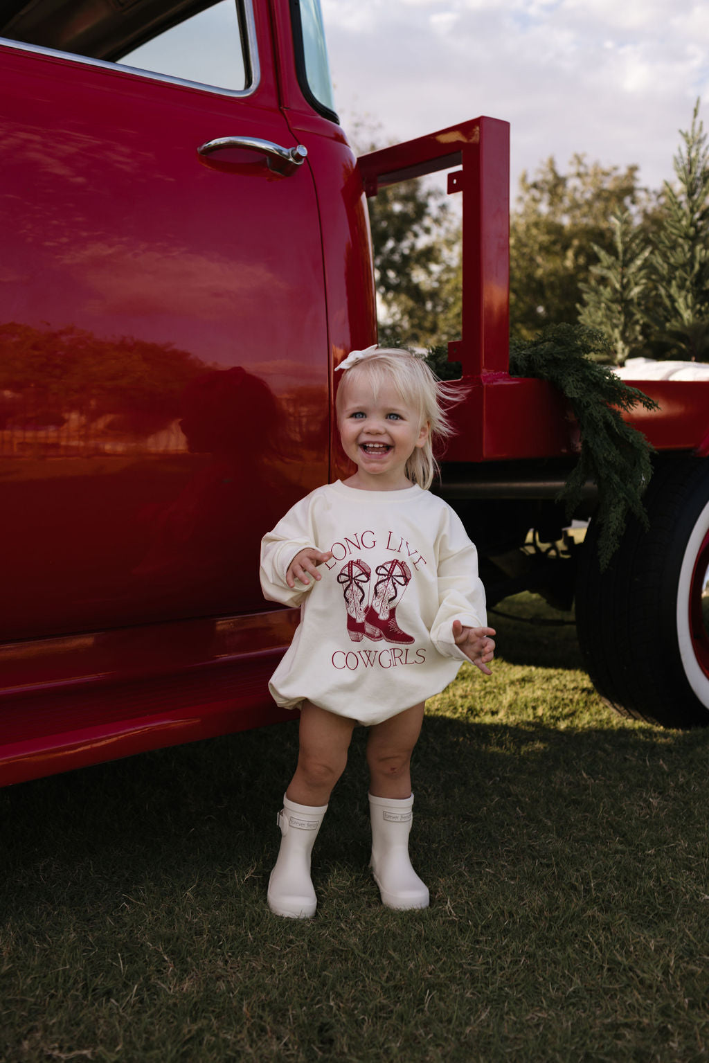 A smiling young child with blonde hair stands beside a red truck, wearing the "Long Live Cowgirls" long sleeve romper by forever french baby and beige boots. Greenery decorates the truck, and a field stretches into the distance, making it perfect for the cooler months.