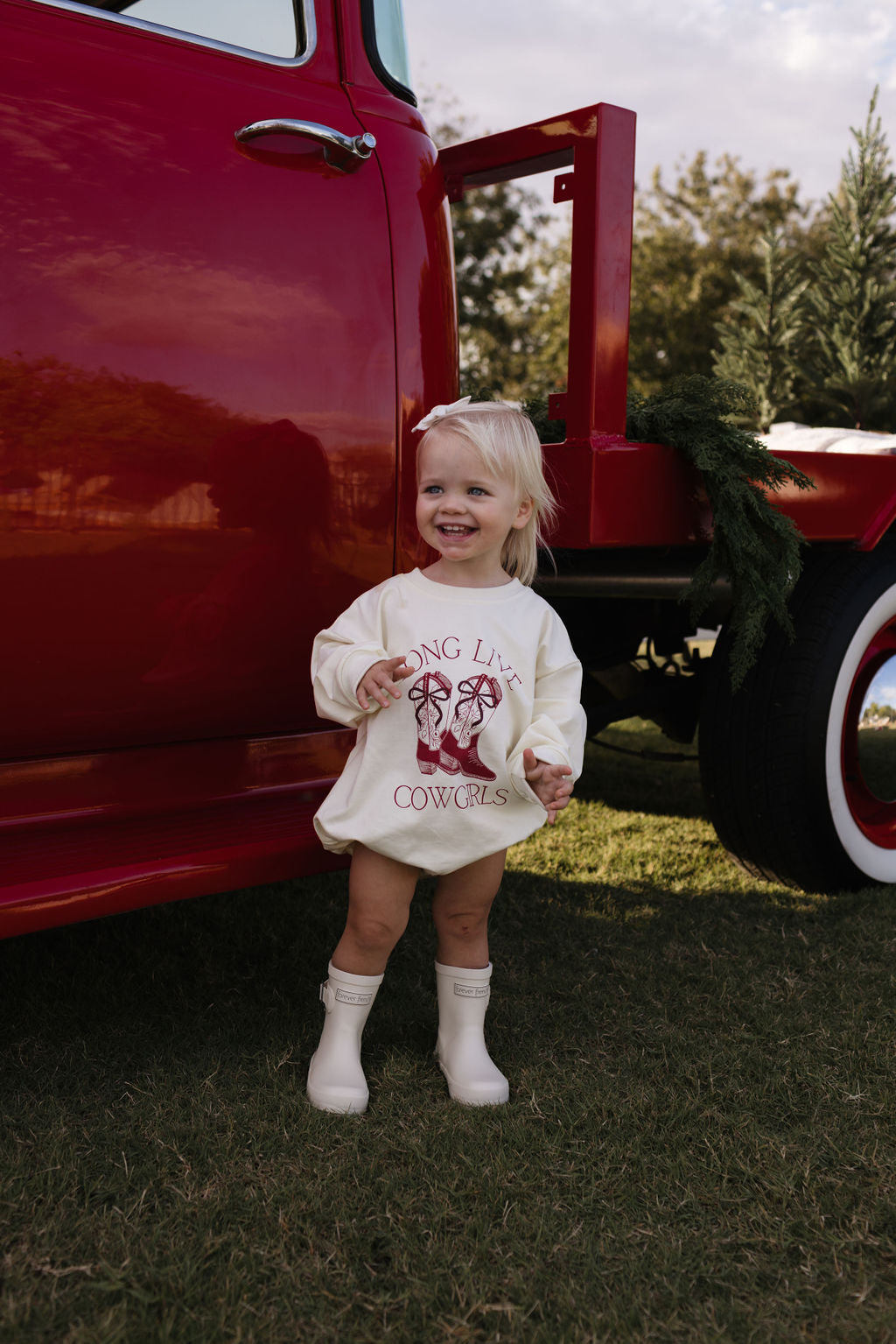 A young child with blonde hair, dressed in a cozy Long Sleeve Romper by forever french baby that reads "Long Live Cowgirls," stands smiling beside a vintage red truck on a grassy area. The truck is adorned with greenery, making it perfect for the cooler months.