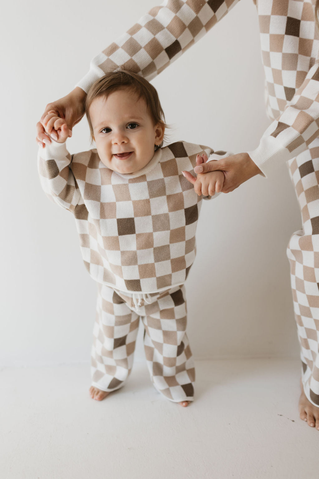 Dressed in the Knit Pant Set | Into the Woods Checkerboard by forever french baby, a toddler joyfully holds hands with an adult. Against a plain white wall, the toddler beams with delight while standing barefoot on the floor.