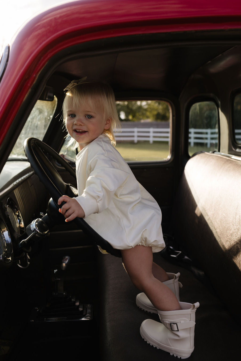 A toddler wearing the "Long Sleeve Romper | Long Live Cowgirls" by forever french baby stands on the seat of a red vehicle, holding the steering wheel. Sunlight pours into the interior, revealing a white fence outside—ideal for adventures during the cooler months.