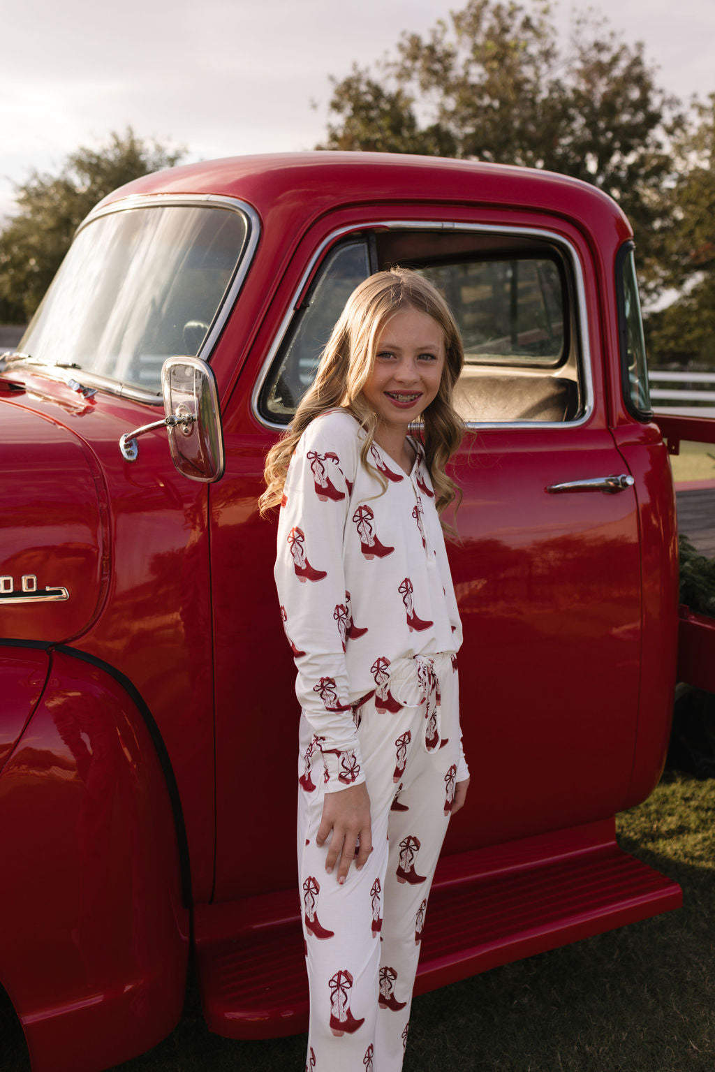 A young girl with long blonde hair stands beside a red vintage truck, wearing cozy Pre-Teen Flare Bamboo Pajamas in the Cowgirl Christmas pattern by lolo webb. The breathable fabric suits the outdoor scene perfectly as she stands on the grassy area with trees in the background and the sun setting softly.