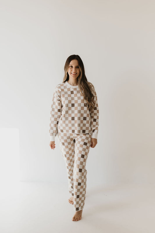 A person with long hair is smiling and walking barefoot in a minimalist setting, dressed in the cozy Women's Knit Pant Set, "Into the Woods Checkerboard," by forever french baby. The background is plain and softly lit.