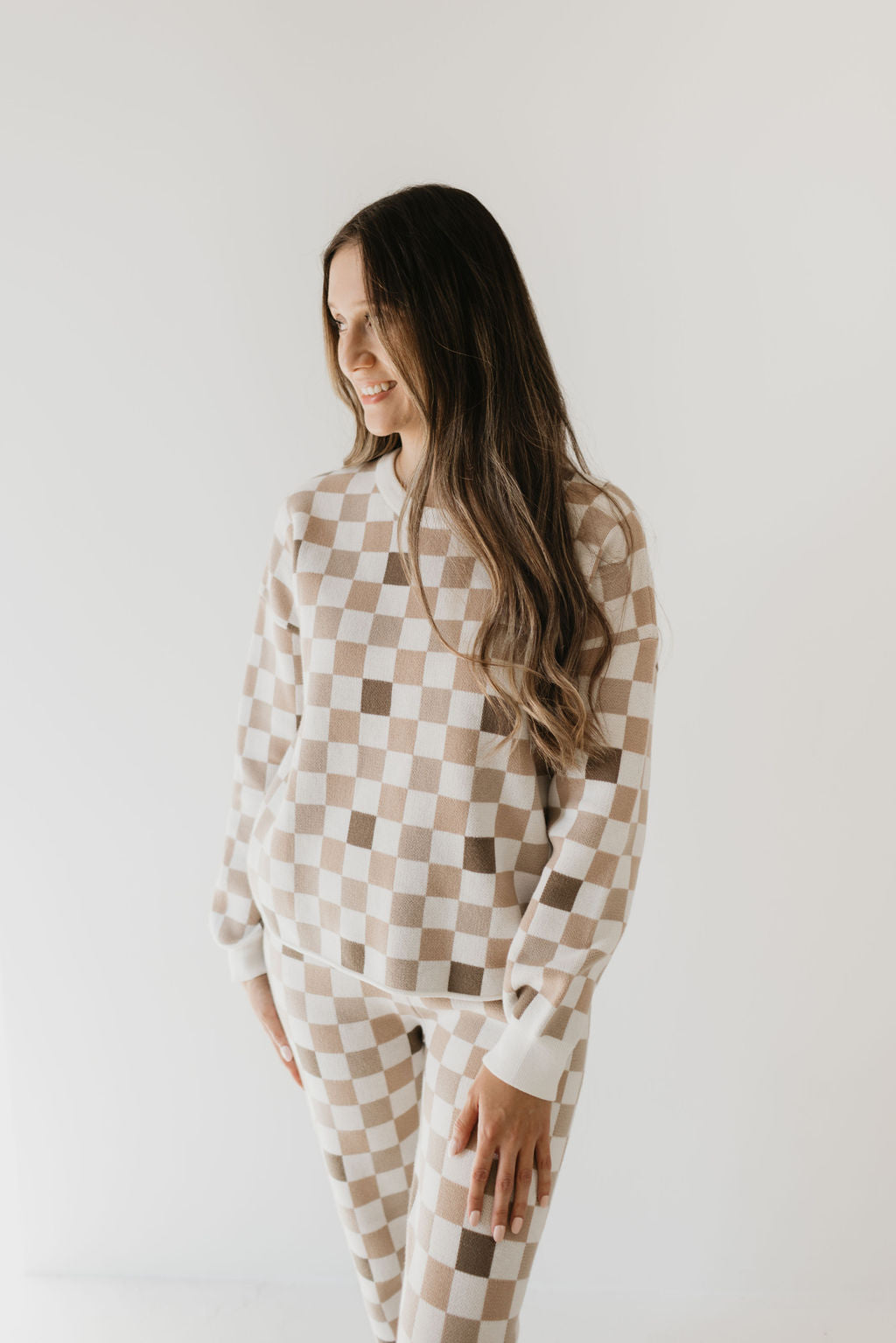 A woman with long hair wears the Women's Knit Pant Set from forever french baby’s Into the Woods Checkerboard line, seamlessly blending beige and white checks into a cozy ensemble as she stands against a plain light background. Made entirely from 100% cotton, she gazes to the side with a slight smile, one hand confidently placed on her hip.