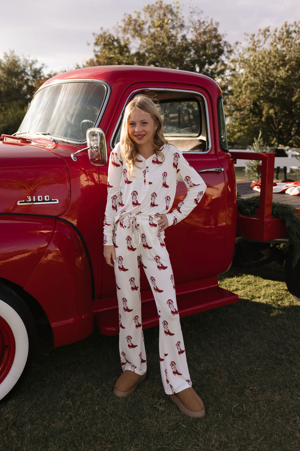 A young girl stands smiling in front of a vintage red truck, dressed in lolo webb's Pre-Teen Flare Bamboo Pajamas in the Cowgirl Christmas pattern. She is outside on grass, surrounded by trees under a partly cloudy sky. These breathable bamboo pajamas ensure she stays comfy and stylish during festive moments.