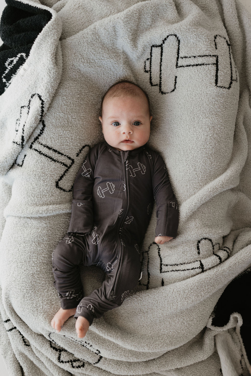 A baby dressed in forever french baby's Bamboo Zip Pajamas in the "Arm Day" style, featuring white dumbbell prints on a dark background, rests comfortably on a plush blanket that matches with similar dumbbell designs. Made from breathable bamboo fabric, the cozy textured material ensures the baby's comfort as they gaze towards the camera.