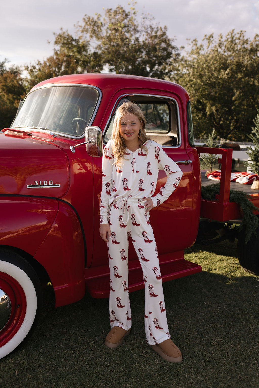 A pre-teen girl, dressed in lolo webb's Pre-Teen Flare Bamboo Pajamas with a Cowgirl Christmas design, stands next to a vintage red truck featuring a wooden bed outdoors. Smiling confidently with one hand on her hip, she is surrounded by trees beneath a partly cloudy sky.