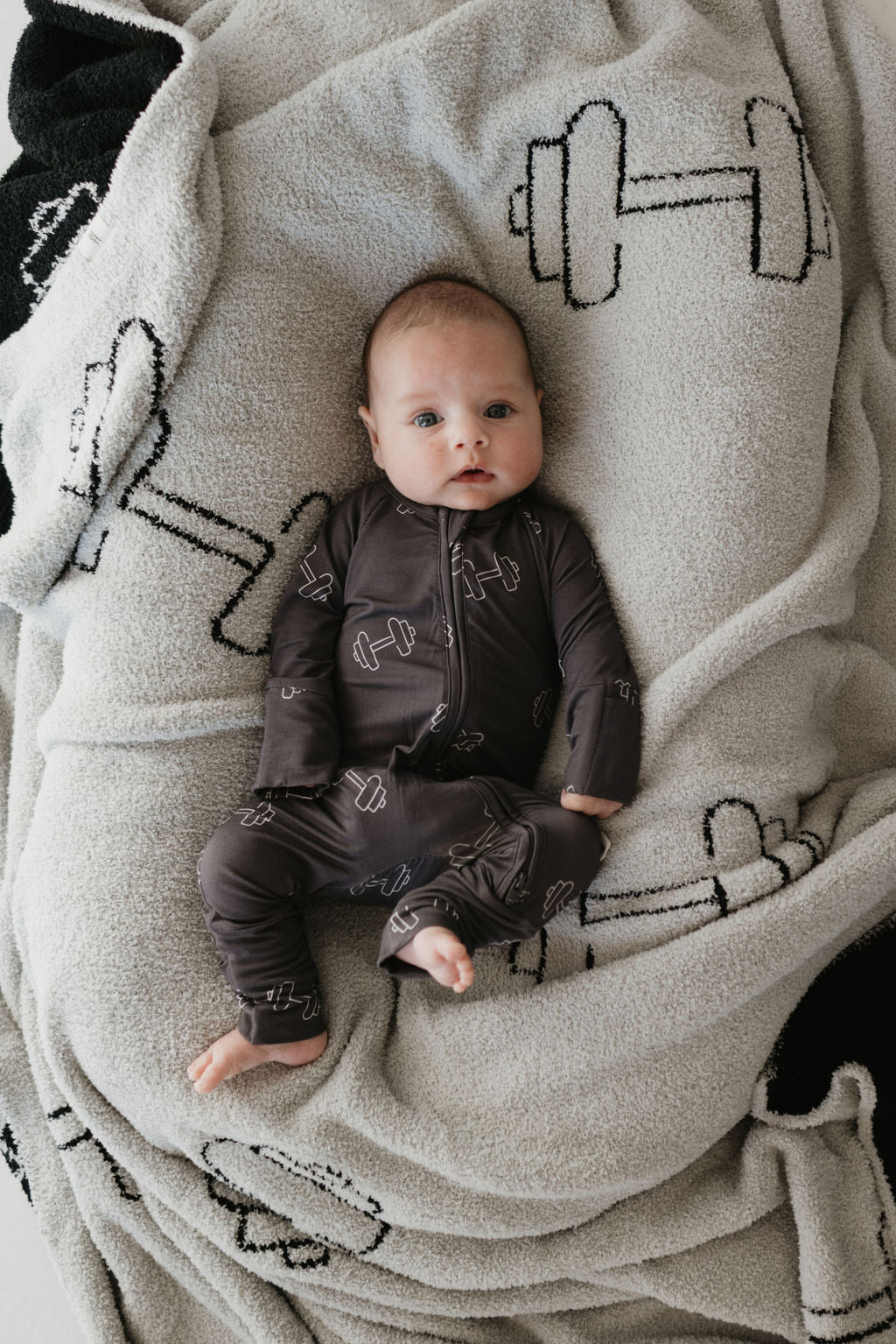 An infant in forever french baby's Bamboo Zip Pajamas in Charcoal Arm Day rests on a white blanket with black dumbbells, gazing at the camera. The breathable onesie showcases a dumbbell pattern and is made from cozy, hypo-allergenic fabric.