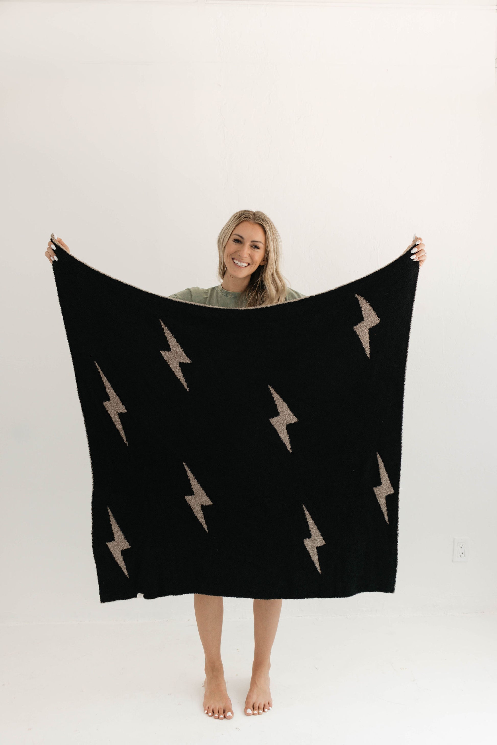 A person standing barefoot on a light-colored floor holds up the Plush Blanket | Brown Lightning Bolt from forever french baby, made from soft microfiber feather yarn. The person is smiling and wearing a green t-shirt. The background is plain and white, highlighting the Oeko-tex certified quality.