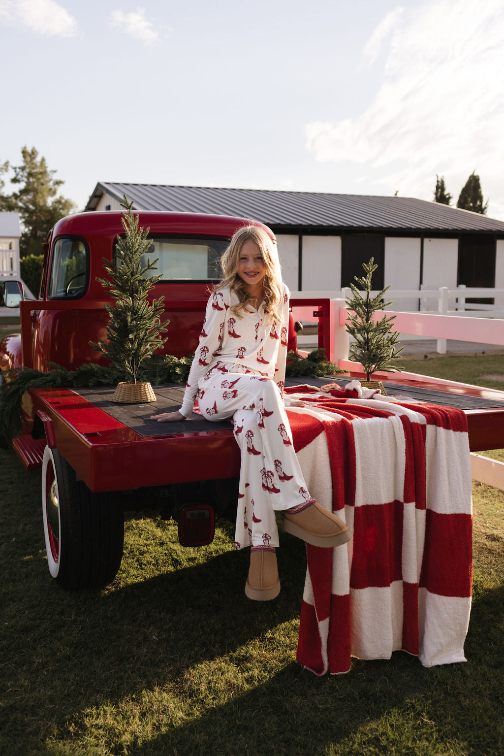 Clad in a pair of lolo webb's Pre-Teen Flare Bamboo Pajamas in the Cowgirl Christmas style, an individual sits on the bed of a red vintage truck. The truck is adorned with small evergreen trees and features a red-and-white checkered blanket. This charming scene takes place outdoors, complete with a white fence and a building visible in the background.