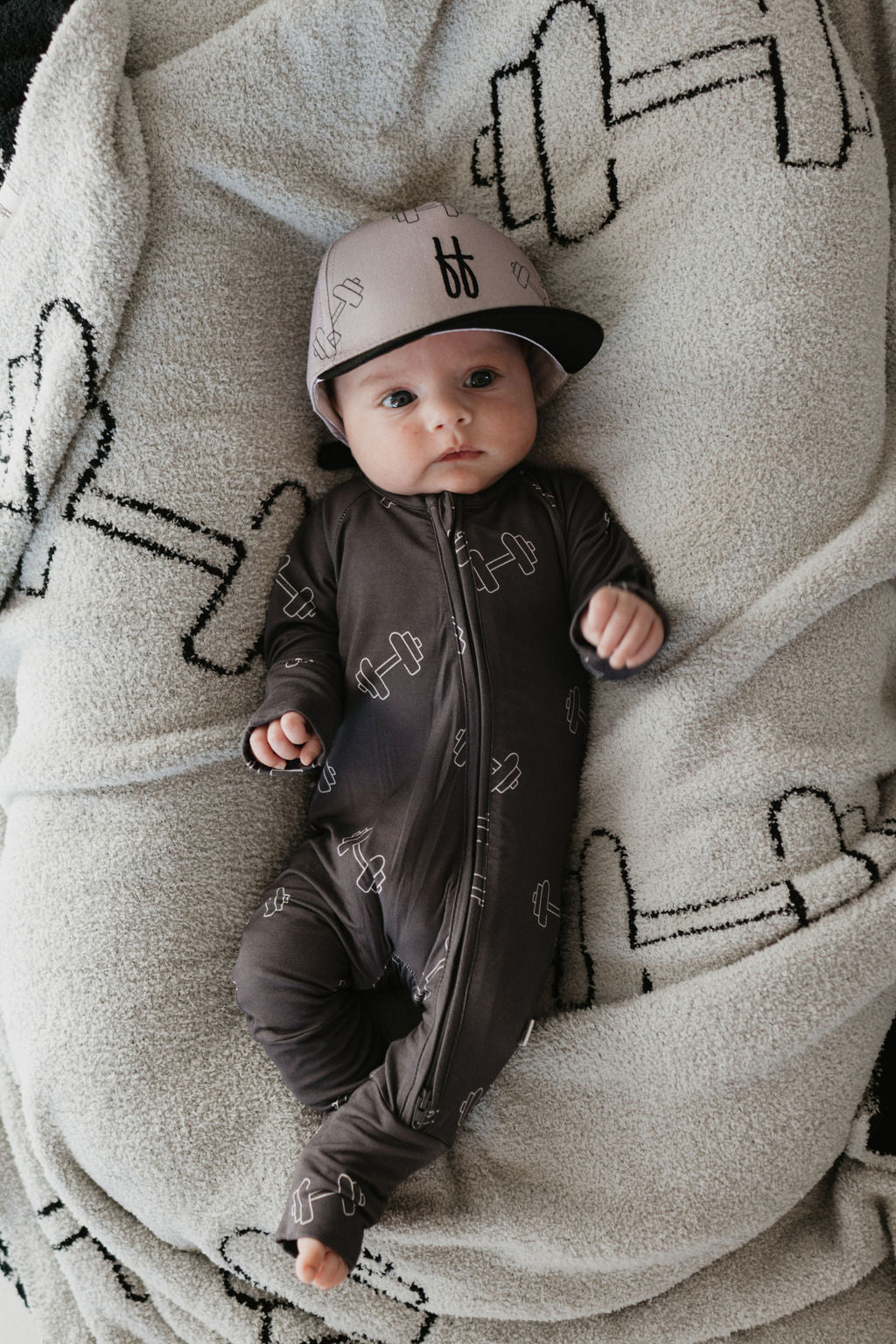 A baby in a Forever French Baby Trucker Hat titled "Arm Day" lies on a gray blanket decorated with dumbbell patterns. The charming design of the trucker hat adds to the scene as the baby gazes up with wide eyes, enjoying both comfort and style.