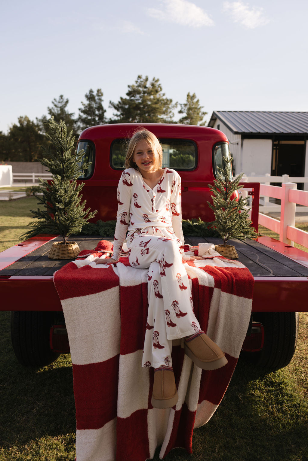 Dressed in lolo webb's Pre-Teen Flare Bamboo Pajamas from the Cowgirl Christmas collection, featuring slippers, a person relaxes on the tailgate of a red vintage truck. The truck bed is decorated with a red and white checkered blanket and two small evergreen trees, with a barn and additional trees visible in the background.