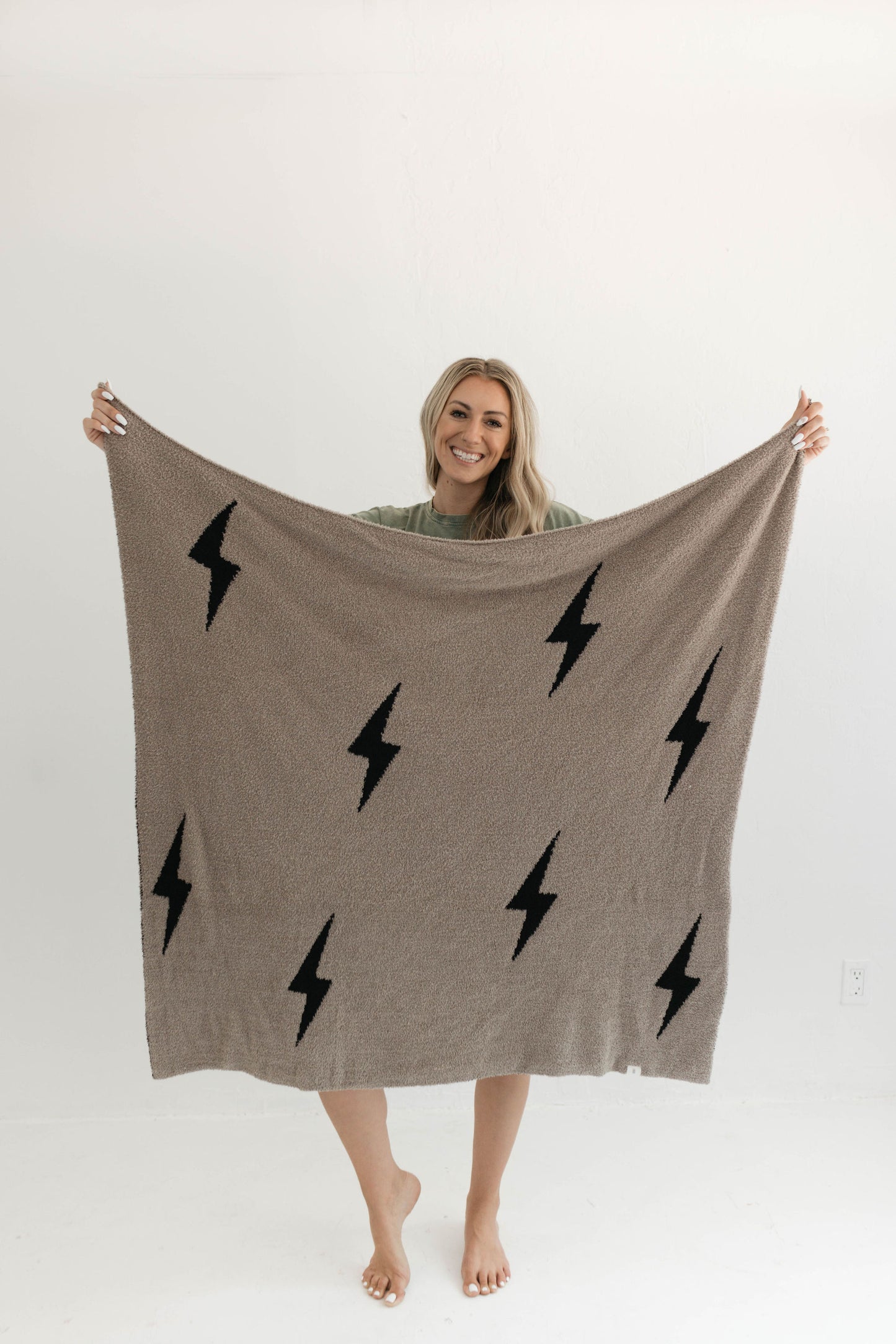 A person with long blonde hair, wearing a green shirt, holds up Forever French Baby's Plush Blanket in Brown Lightning Bolt pattern, made from microfiber feather yarn. The person is smiling and standing barefoot on a plain white background.