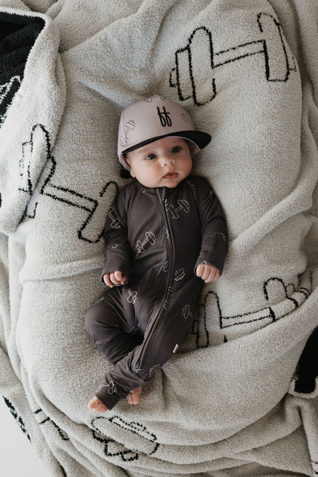 A baby in forever french baby's Bamboo Zip Pajamas | Charcoal Arm Day lies on a blanket with black dumbbells. The cozy, hypo-allergenic bamboo fabric ensures a breathable and coordinated look with the matching cap for ultimate comfort.