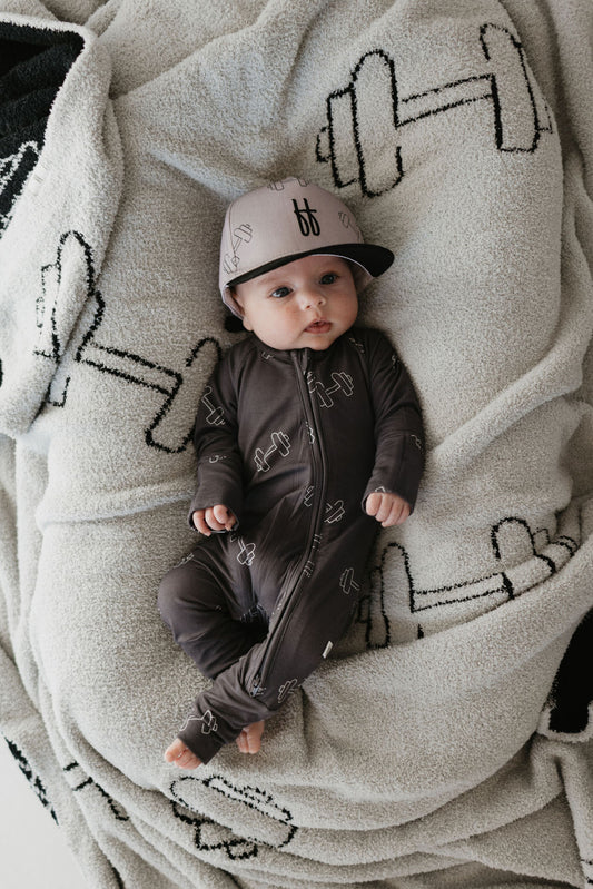 A baby lies on a blanket adorned with dumbbell patterns, wearing a matching dark onesie and the Children's Trucker Hat | Arm Day. The hat provides sun protection with its logo, while the onesie features a matching dumbbell design. Styled in Forever French Baby fashion, this little one sets the trend for chic comfort.