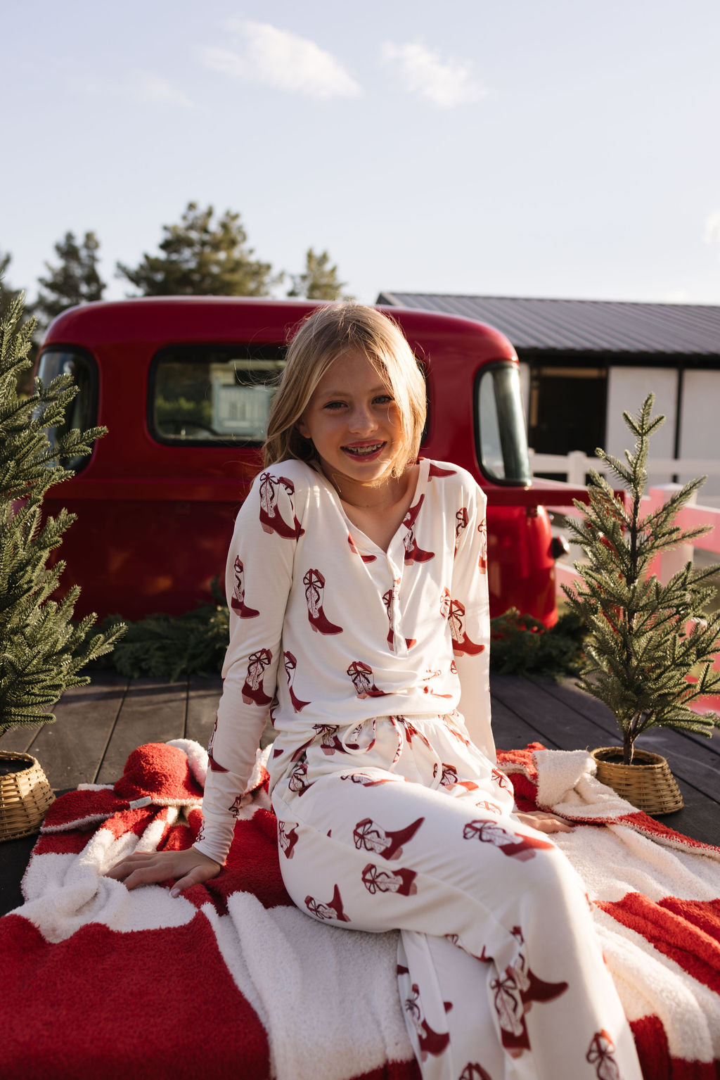 A smiling child sits on a red and white blanket outdoors, surrounded by small pine trees. They're wearing festive Pre-Teen Flare Bamboo Pajamas in the Cowgirl Christmas design from lolo webb, known for their breathable fabric. A vintage red truck is in the background under a clear, sunny sky.