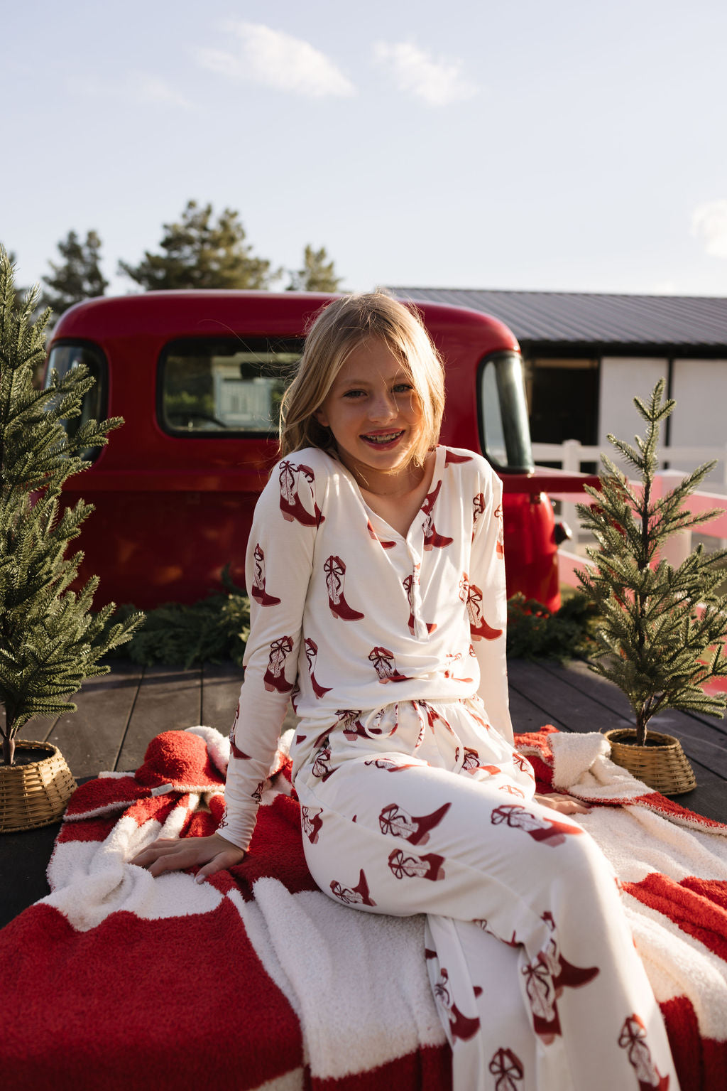 A young person wearing lolo webb's Pre-Teen Flare Bamboo Pajamas in the Cowgirl Christmas pattern sits on a breathable fabric blanket outdoors, surrounded by small pine trees. A red vehicle is parked in the background, and a building is visible under a clear sky.