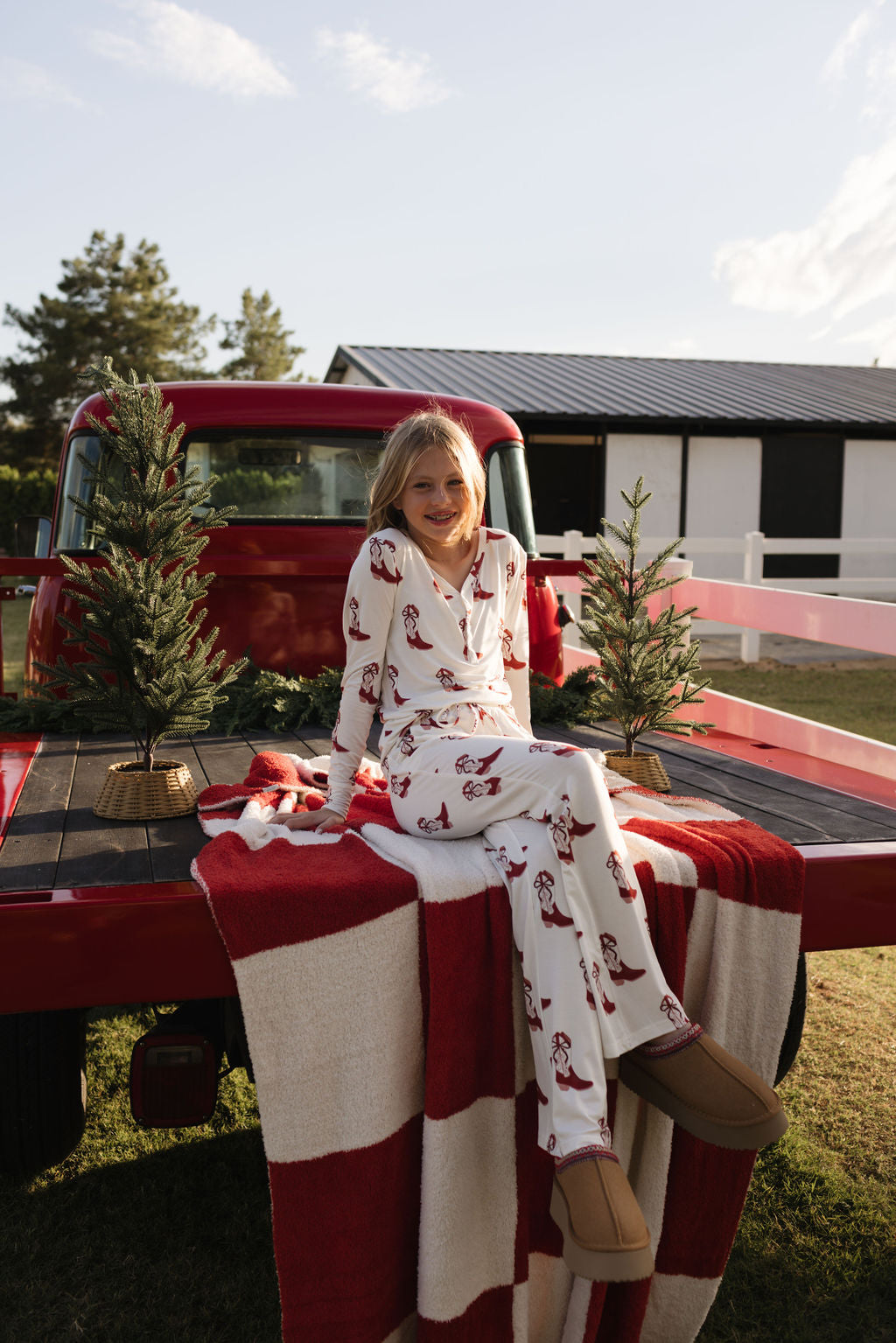 A child sits on the tailgate of a red truck, decorated with small Christmas trees and a cozy Plush Blanket | the Quinn by lolo webb, which is Oeko-tex certified in red-and-white. Dressed in festive pajamas and smiling broadly, the scene is set against a picturesque barn and trees beneath a clear sky.