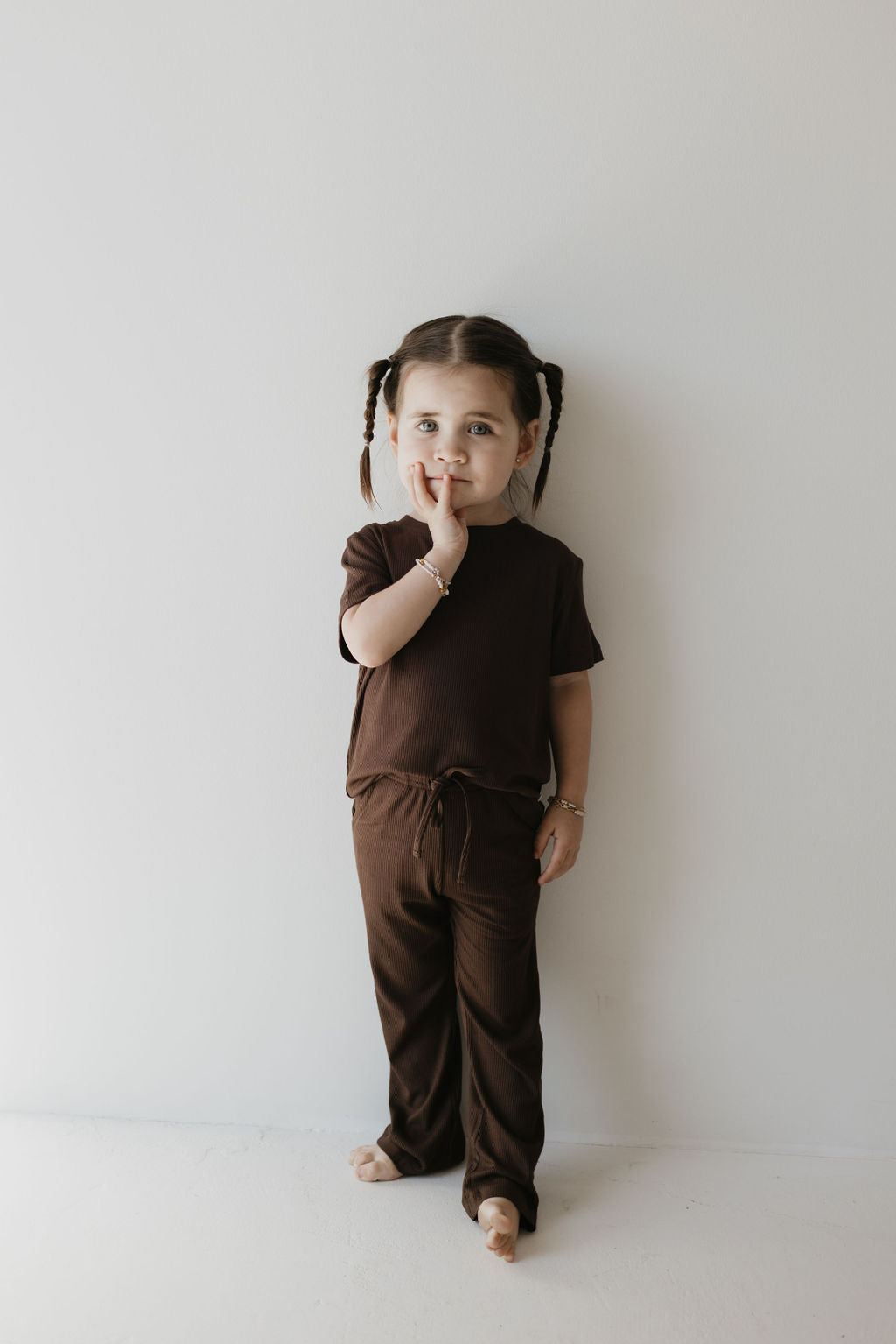 A young child with braided hair, dressed in the "Toddler Short Sleeve Bamboo Lounge Set | Coffee Bean" by forever french baby, stands barefoot against a light wall, holding a finger to their lips.