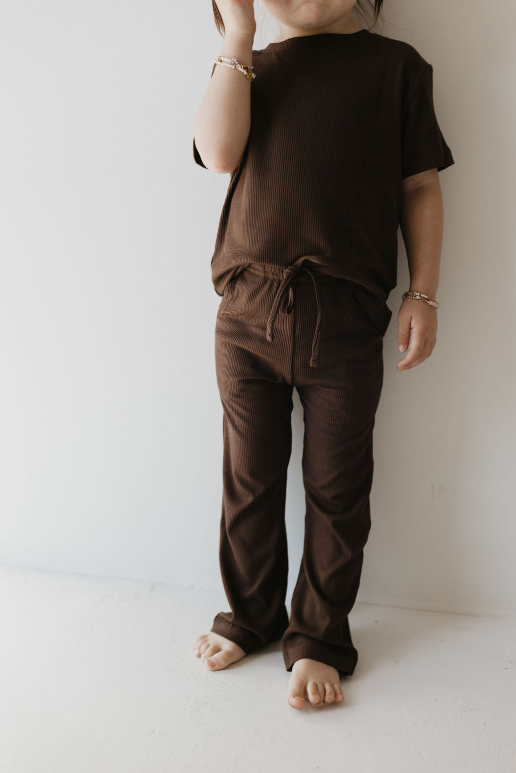 A child stands barefoot against a white wall, dressed in the Toddler Short Sleeve Bamboo Lounge Set in Coffee Bean from forever french baby. The short-sleeved top and matching drawstring pants crafted from breathable bamboo fabric provide comfort and style. Touching their face with one hand, the child's bracelets enhance the charm of this hypo-allergenic ensemble.