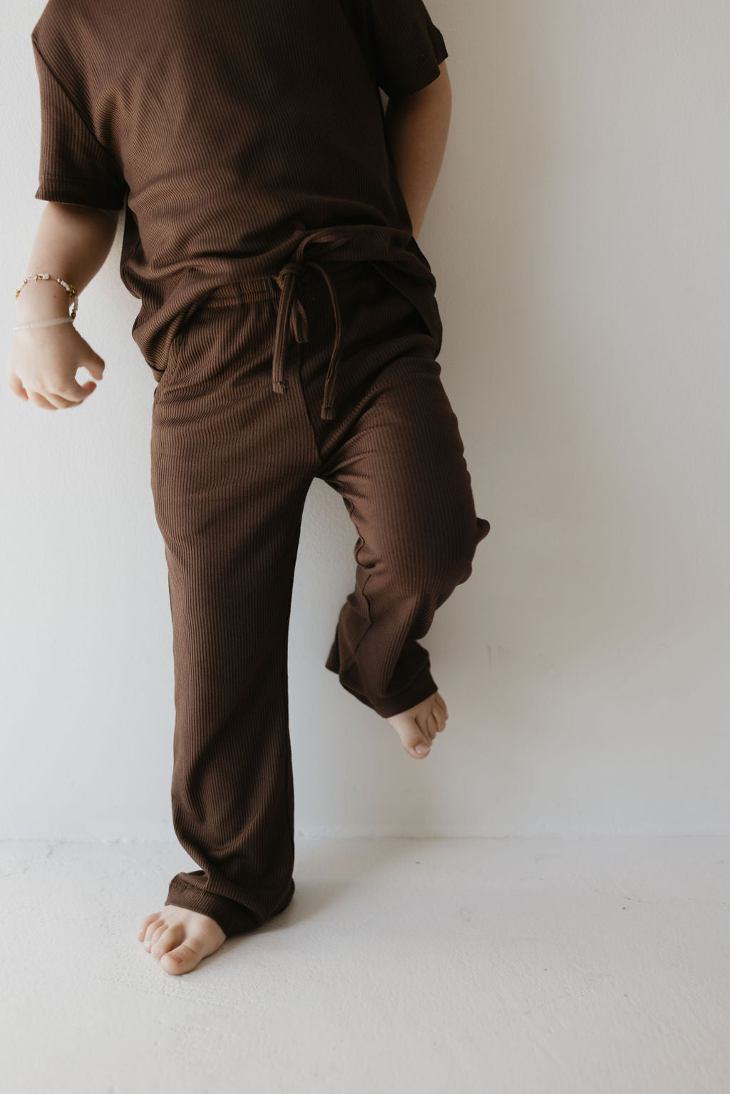 A child wearing the Toddler Short Sleeve Bamboo Lounge Set in Coffee Bean from forever french baby balances on one leg against a plain white wall. Barefoot and relaxed, the child has one hand partially visible. This hypo-allergenic outfit features a short-sleeve top and matching pants tied at the waist, made from breathable bamboo fabric.