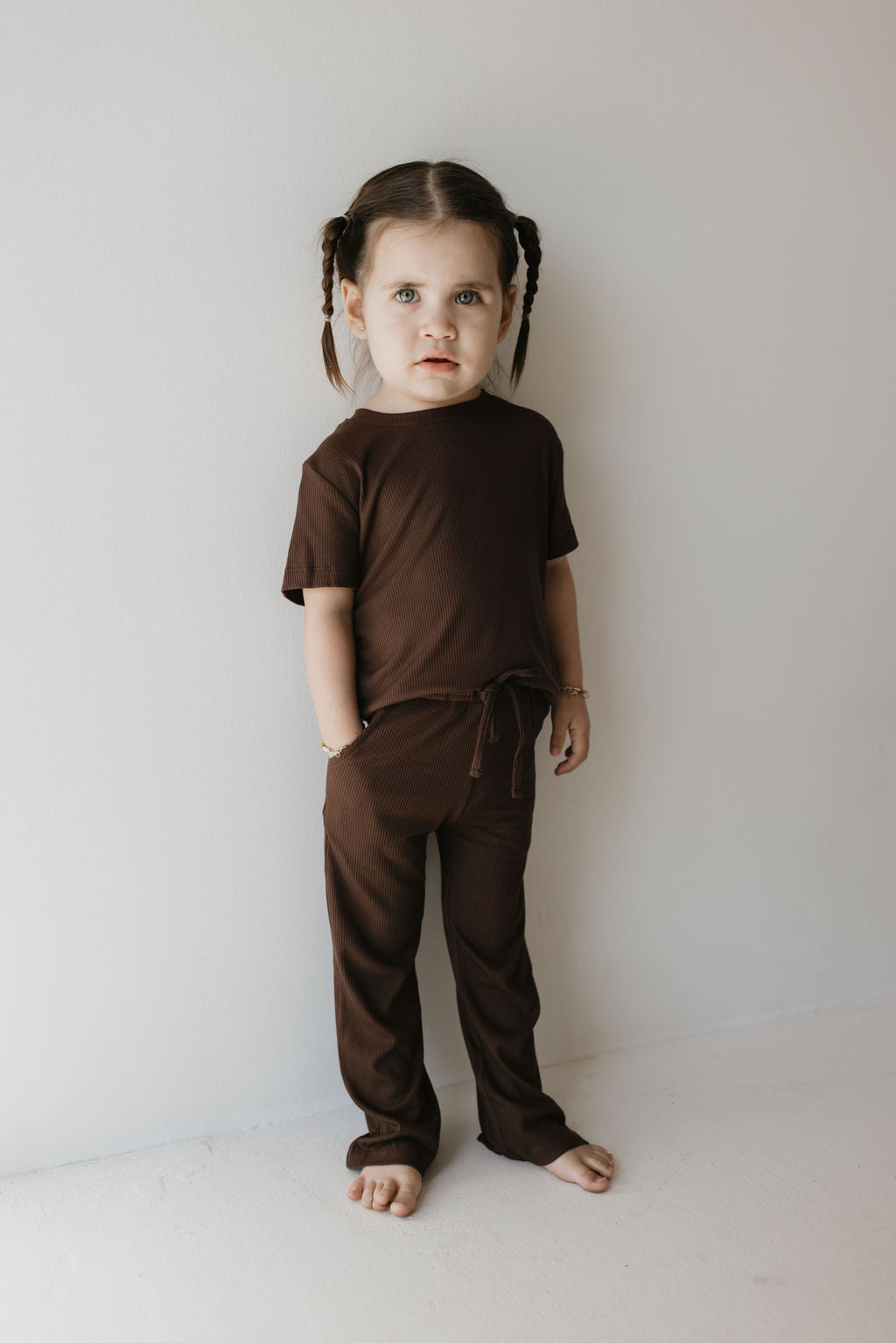 A young child with brown braided hair stands against a light-colored wall, dressed in the forever french baby Toddler Short Sleeve Bamboo Lounge Set in Coffee Bean. The breathable outfit is made from hypo-allergenic bamboo. The child is barefoot with hands in pockets, gazing at the camera with a neutral expression.