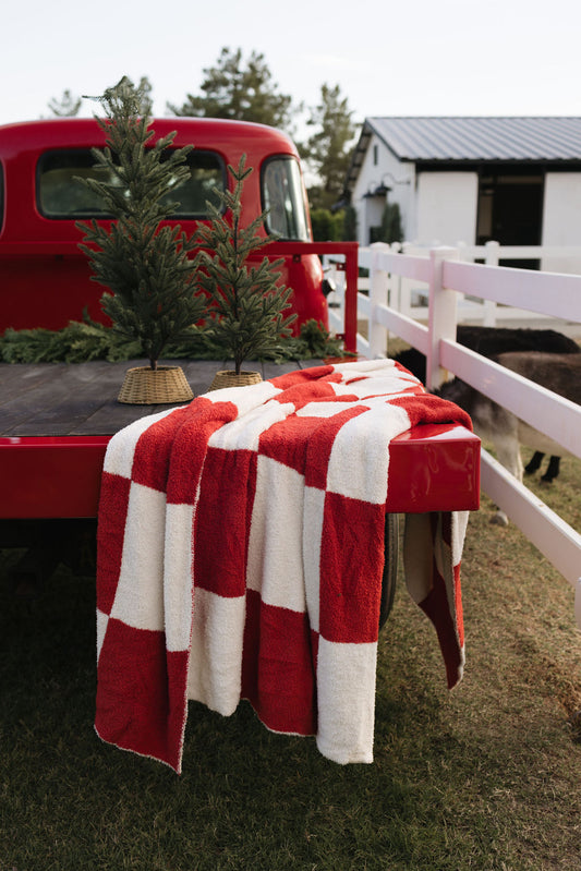 A vintage red truck with a lolo webb Plush Blanket | the Quinn, crafted from Oeko-Tex certified materials, draped over the tailgate. Two small evergreen trees sit in the truck bed. The scene is set against a white fence, with a barn visible in the background.​