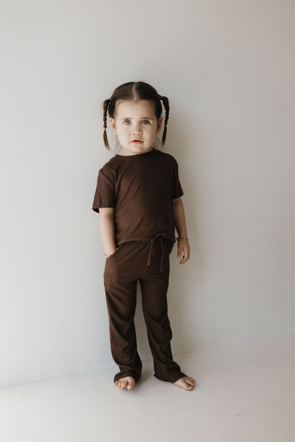 A young child with pigtails stands barefoot against a light-colored wall, wearing the Toddler Short Sleeve Bamboo Lounge Set in Coffee Bean from forever french baby. They gaze directly at the camera with a neutral expression.