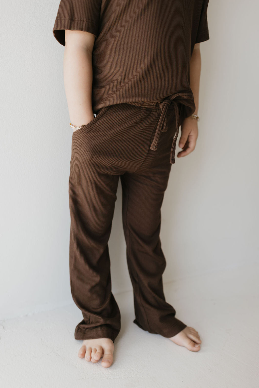 A person stands against a light-colored wall, dressed in the forever french baby Toddler Short Sleeve Bamboo Lounge Set in Coffee Bean. Their hands are tucked into the pockets, and their bare feet rest on the white floor. Their face is not visible.