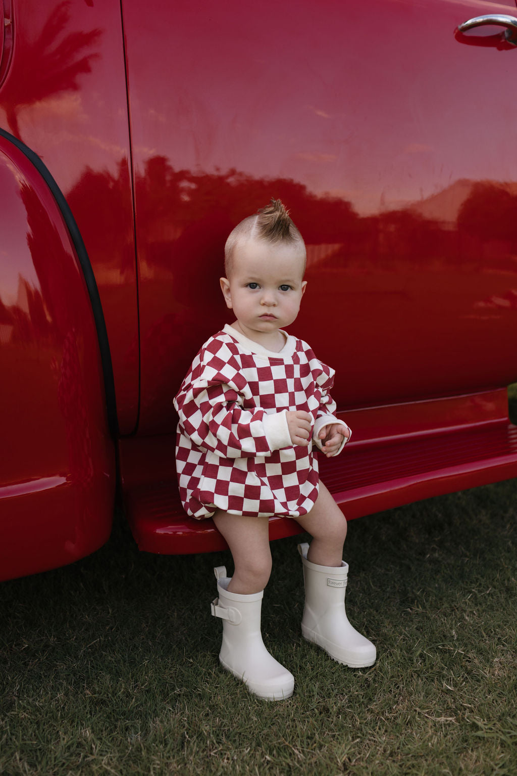 A baby with a fashionable mohawk is seated on the side step of a red vehicle, charmingly dressed in a checkered red and white Long Sleeve Romper by lolo webb, known as the Quinn, paired with snug white boots. The little one, aged newborn to 24 months, gazes curiously at the camera while grass sways softly in the background.