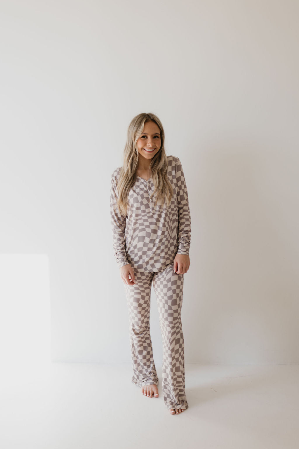 A woman with long blonde hair is smiling while wearing the Women's Bamboo Pajamas in the Smokey Wave design by forever french baby. The breathable fabric ensures her comfort as she stands barefoot against a plain white background, showcasing the pajamas' elegant design.