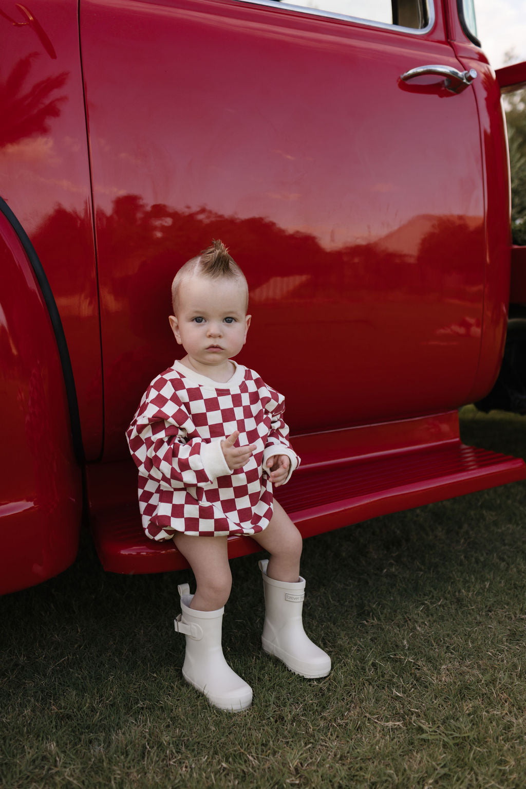 Dressed in a long sleeve romper from lolo webb's "the Quinn" collection, featuring a red and white checkered design, the baby sits on the step of a vibrant red vehicle. Sporting stylish white boots and a charming mohawk hairstyle, the little one is all set for cooler weather. The lush green surroundings add an extra layer of charm to this delightful scene.