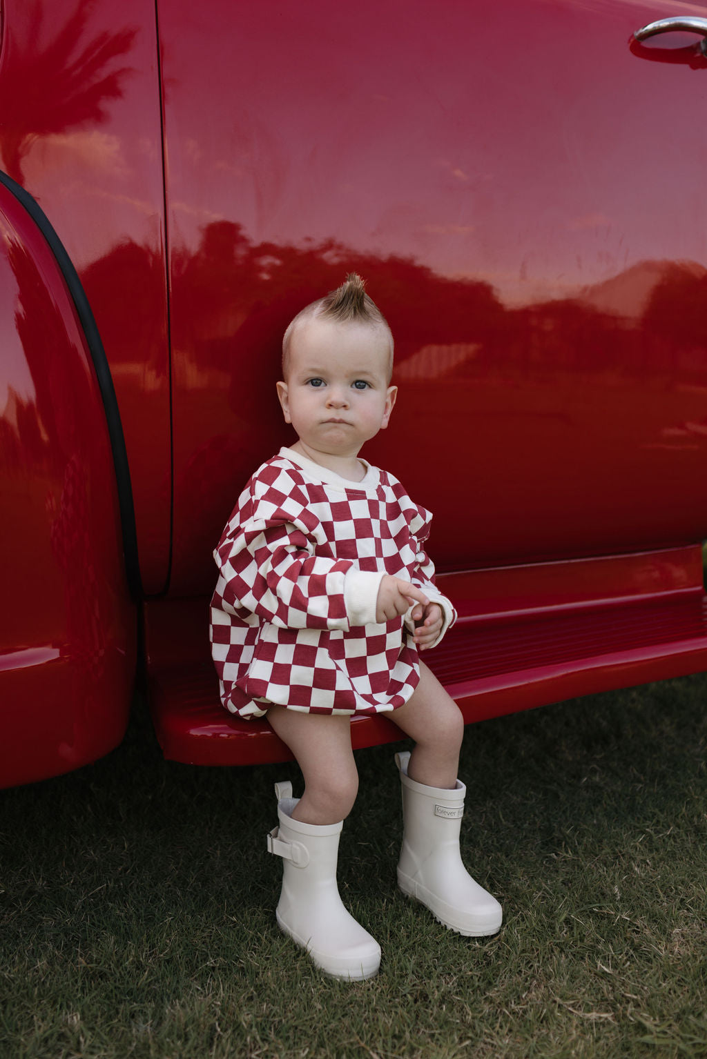 A baby with a mohawk hairstyle is seated on the step of a red vehicle, wearing the cozy Long Sleeve Romper from lolo webb's "the Quinn" collection. The playful red and white checkered design paired with white boots is perfect for cooler months as they gaze directly at the camera while sitting on the grass.