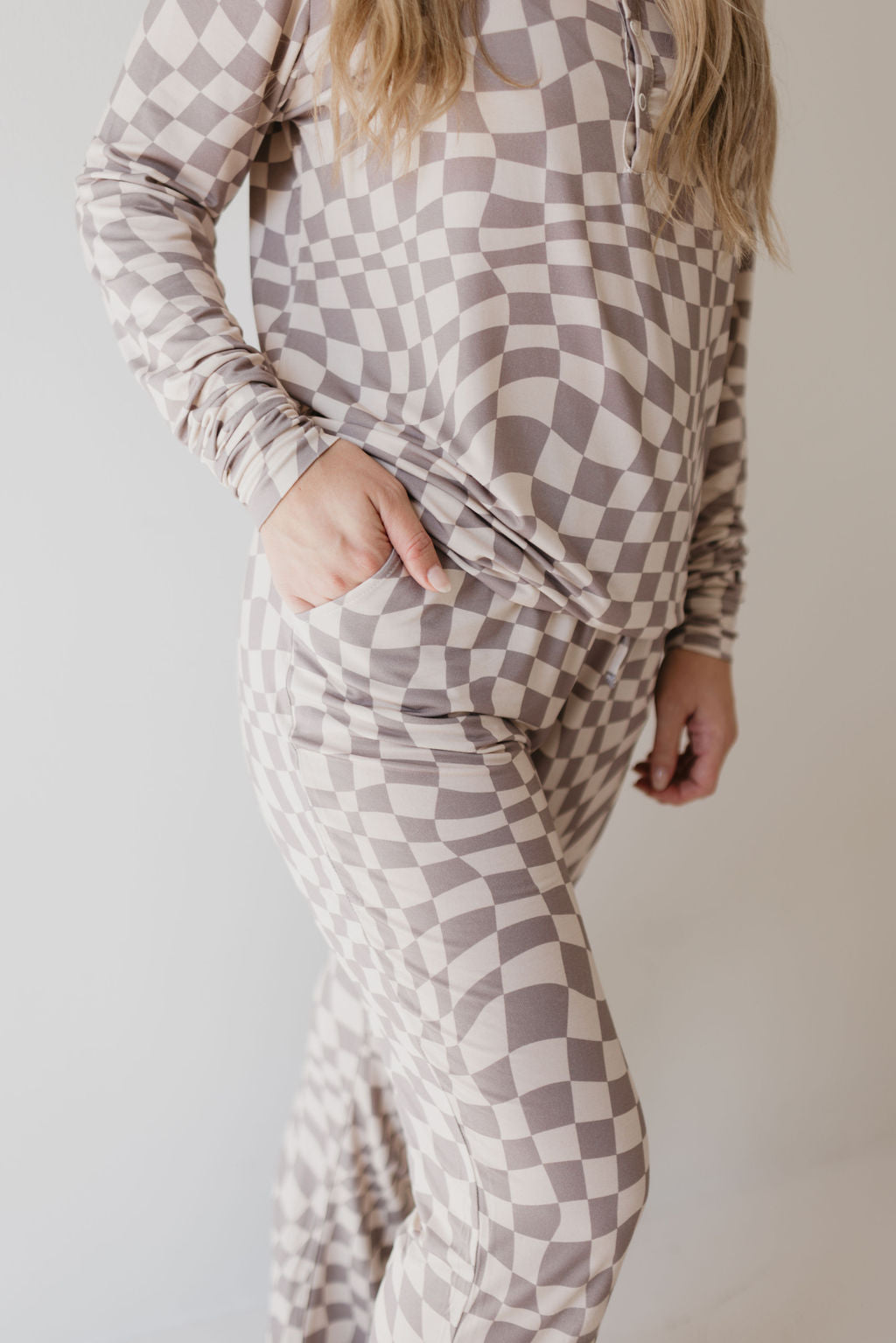 A person is dressed in the Women's Bamboo Pajamas in Smokey Wave, featuring matching geometric patterns in neutral tones from the brand forever french baby. The image highlights the torso and legs, with a hand casually placed in one of the pants pockets. This breathable attire contrasts beautifully against a plain, light-colored background.