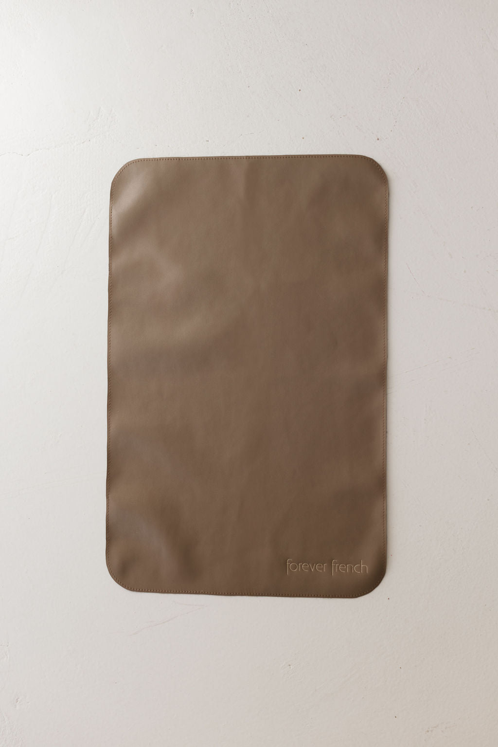 A rectangular taupe vegan leather placemat with rounded corners lies flat on a light textured surface, showcasing the embossed text "forever french baby" in the bottom right corner.