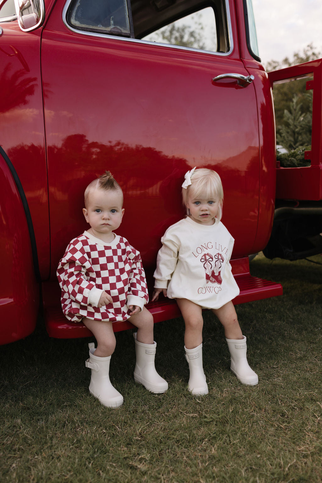 Two young children sit on the step of a red vintage truck, dressed in checkered and printed outfits perfect for cooler months. With white boots and a tree in the background, they enjoy the outdoors. The Long Sleeve Romper by lolo webb, known as the Quinn, is ideal for newborns to 24 months joining their playful adventures.