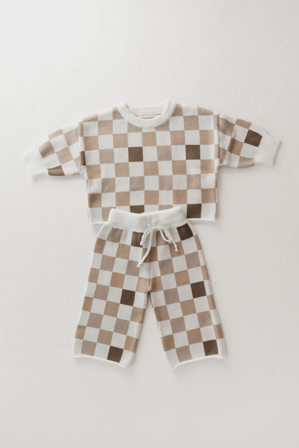 The "Into the Woods Checkerboard" Knit Pant Set by forever french baby is a children's two-piece outfit made from 100% cotton. It features a brown and white checkerboard design and includes an oversized long-sleeve top paired with drawstring pants, both displayed flat against a white background.