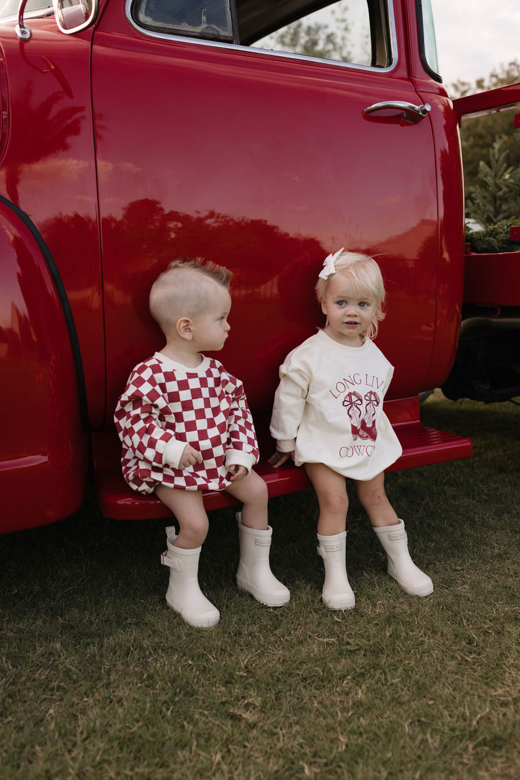 Two toddlers, both in the newborn to 24 months range, sit on the step of a vintage red truck. One is dressed in a checkered outfit with rain boots, while the other wears a white "Long Sleeve Romper | the Quinn" from lolo webb that has "Long Live Cowboys" emblazoned on it. They’re nestled on green grass amid trees, making it an ideal setting for cooler months.