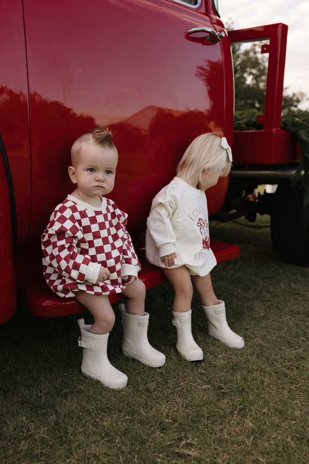 Two toddlers sit on the side step of a vintage red truck. The child on the left wears a red and white checkered outfit, while the other is dressed in a cozy Long Sleeve Romper from lolo webb's Quinn collection, ideal for cooler months. Both are seated on grass with trees in the background, creating a charming scene suitable for ages newborn to 24 months.