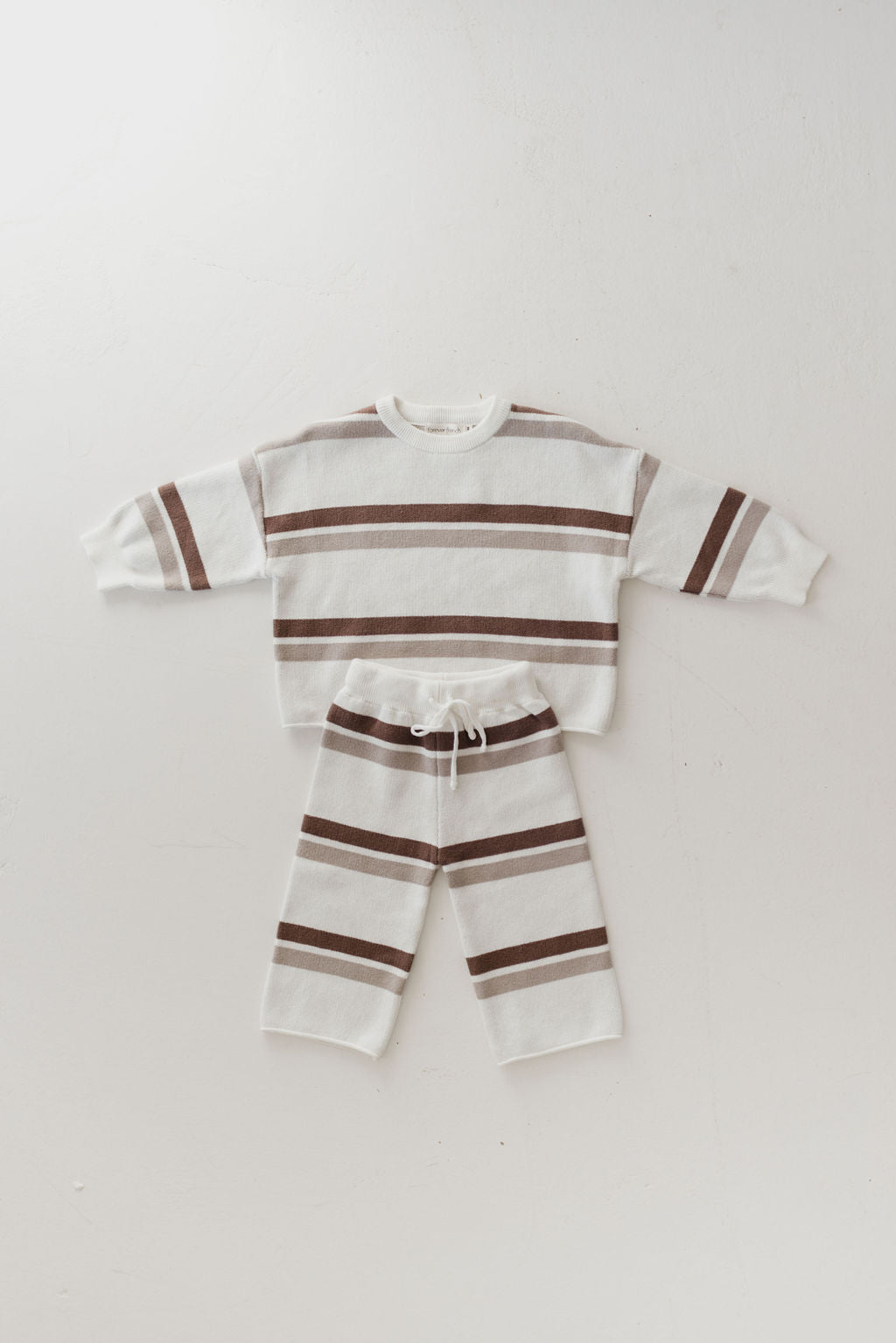 The Woodland Stripe Knit Pant Set by forever french baby is an ideal two-piece outfit for toddlers' fall wardrobes. This laid-flat, 100% cotton knit set includes a long-sleeve sweatshirt and pants, both adorned with wide horizontal stripes in white and brown. The pants come with a drawstring waist to ensure comfort.