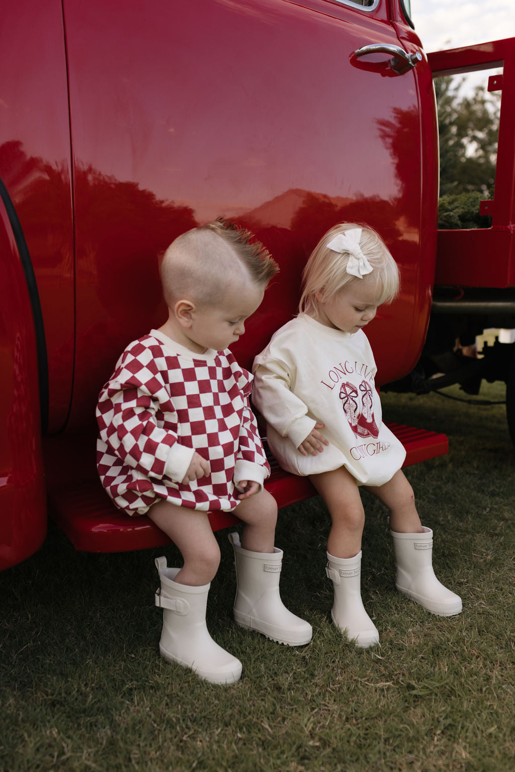 Two toddlers, bundled for the cooler months, sit on a red truck step. One wears a red and white checkered outfit, while the other is dressed in lolo webb's Long Sleeve Romper | the Quinn. Both have short blonde hair and wear white rain boots. Grass and trees create a picturesque backdrop.