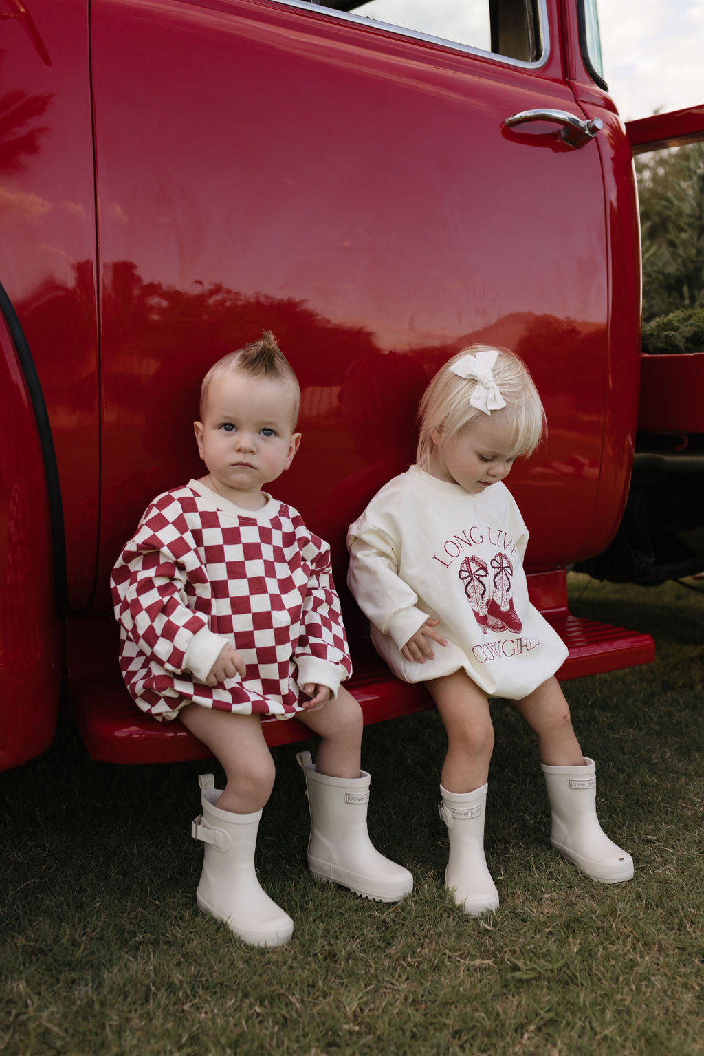 Two toddlers, impeccably dressed for the cooler months, are sitting on the step of a vintage red vehicle. One child is wearing the Quinn Long Sleeve Romper by lolo webb in a red and white checkered pattern, and the other is dressed in a cream outfit with matching boots. The child in cream adorns a bow in their hair. Both children wear stylish white boots, perfect for ages newborn to 24 months.