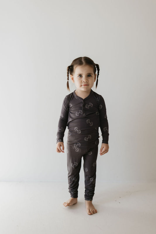 A young child with braided hair stands against a plain background wearing forever french baby's Bamboo Two Piece Pajamas in Charcoal Arm Day. This breathable set complements the child's neutral expression as they gaze at the camera.