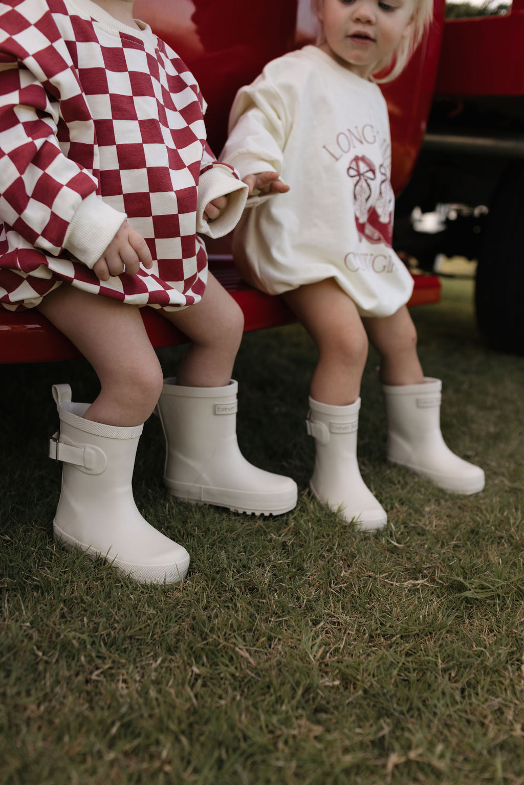 Two young children sit on a red bench, their white rubber boots gleaming. One child sports a red and white checkered outfit from the lolo webb collection, while the other wears the Long Sleeve Romper | the Quinn in cream with a red design—an ideal choice for cooler months. They're enjoying the fresh air in an outdoor setting designed for newborns to 24 months.